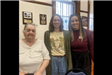 A senior stands with her hands on the back of a chair, along with two smiling young women.